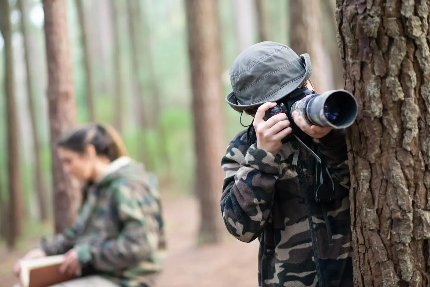 Konzentrierter Junge mit Kamera im Wald. Schuljunge im Mantel und mit Panamahut, der fotografiert. Verschwommene Mutter im Hintergrund. Konzept: Kindheit, Natur, Freizeit.